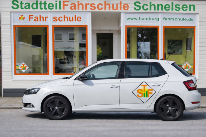 Weißes Auto mit Logo der Fahrschule vor einem Gebäude mit orangefarbenen Fenstern.